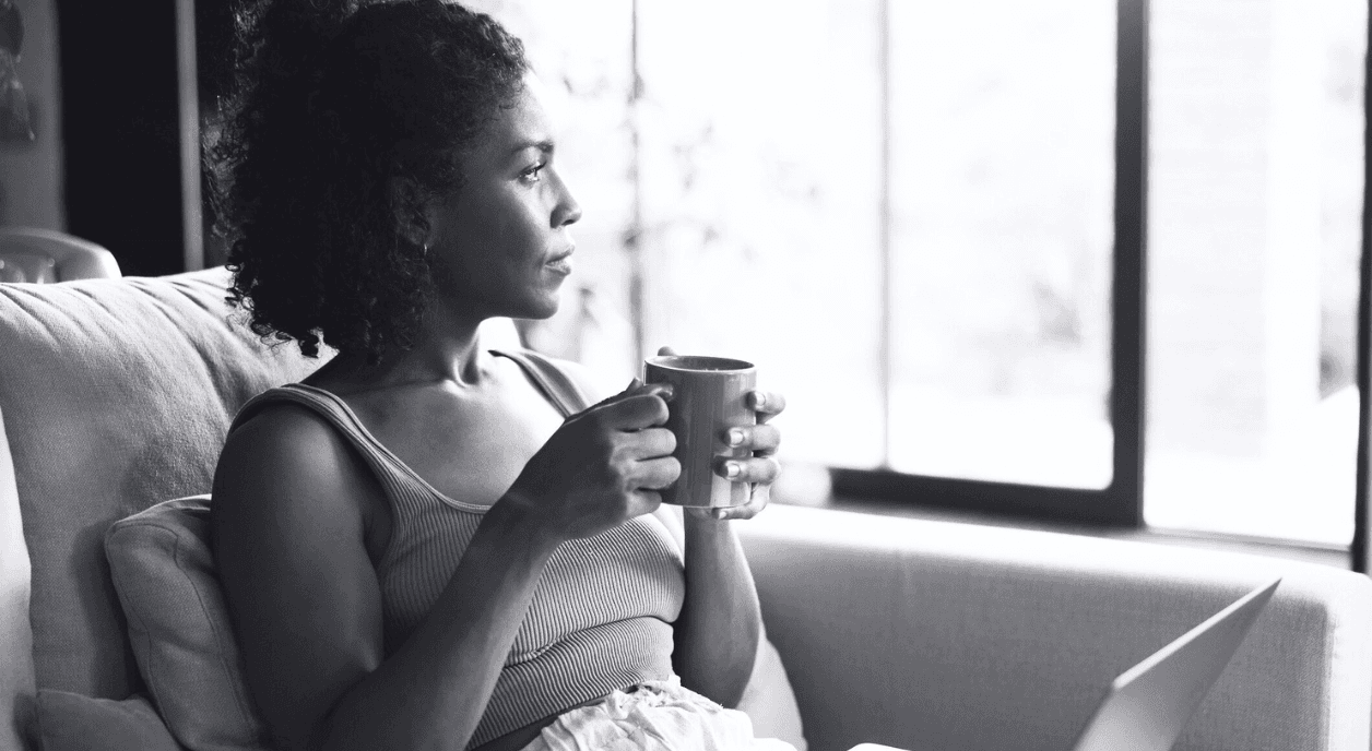 A Black woman on her couch drinking coffee with her laptop open.